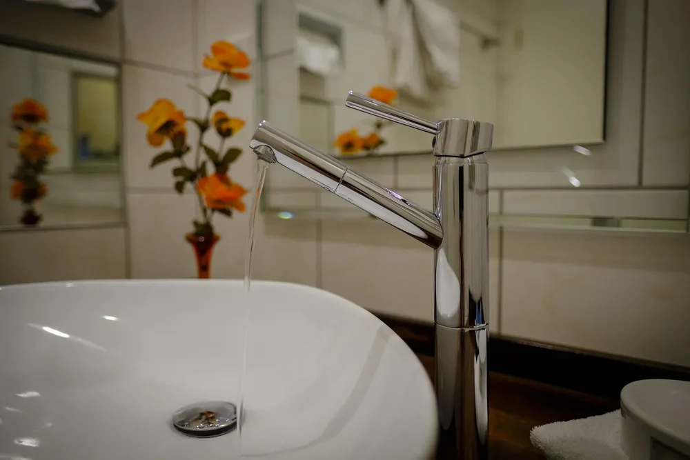 Close-up of a chrome bathroom faucet with water flowing into a white sink, orange flowers blurred in the background.