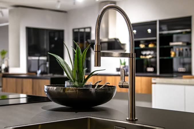 Brushed stainless steel high-arch kitchen faucet over a sink with a dark countertop and a black ceramic planter containing a green plant on the island.